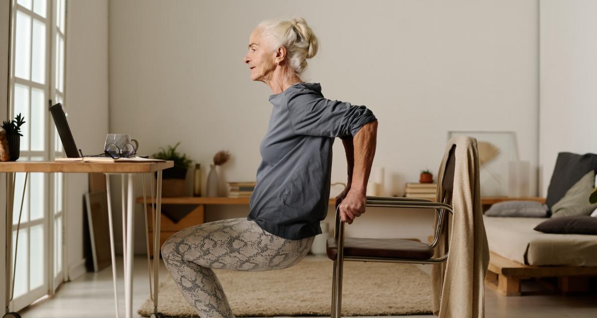 Une femme âgée fait des squats assistés avec une chaise.