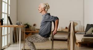 Yoga senior : bienfaits, conseils et postures recommandées - Une femme âgée fait du yoga chez elle (la posture de l'arbre) en s'aidant d'une chaise.