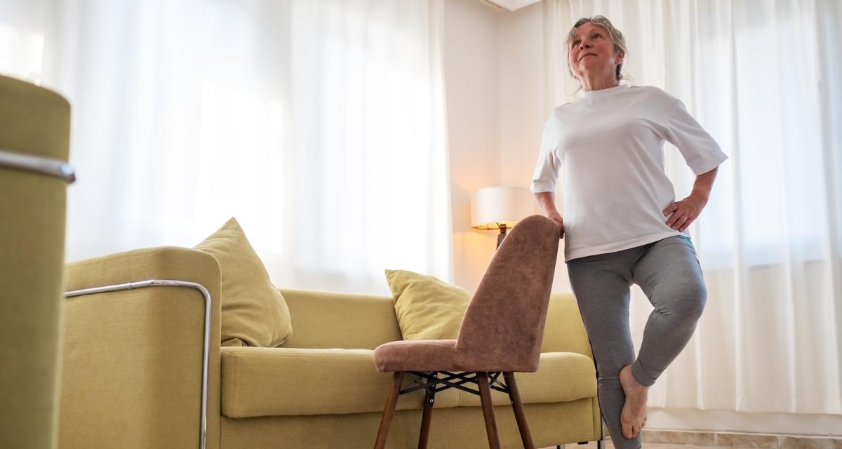 Une femme âgée fait du yoga chez elle (la posture de l'arbre) en s'aidant d'une chaise.